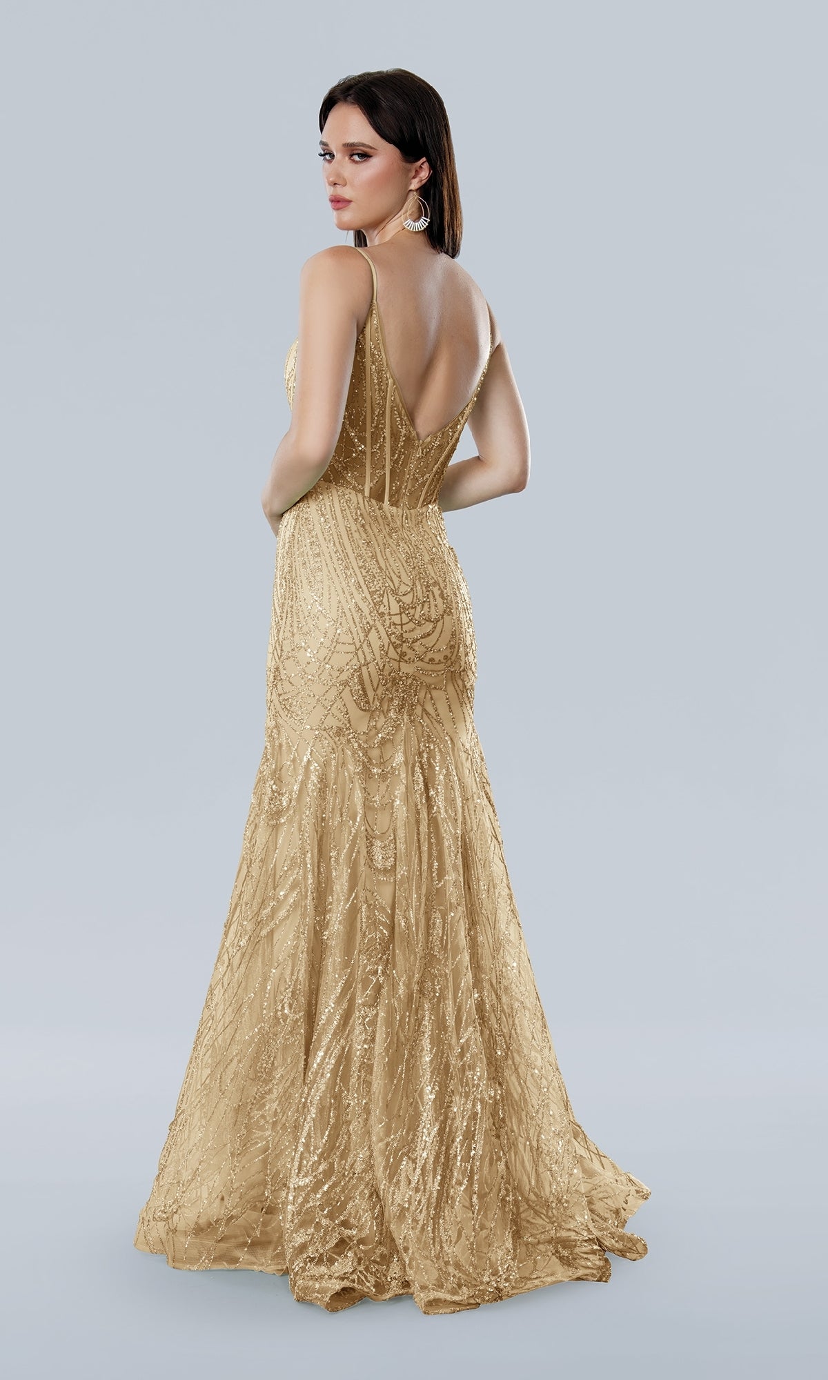 A woman stands against a light gray background, looking over her shoulder in the Stella Couture 25105—a floor-length, sleeveless gold glitter prom dress with a low back.